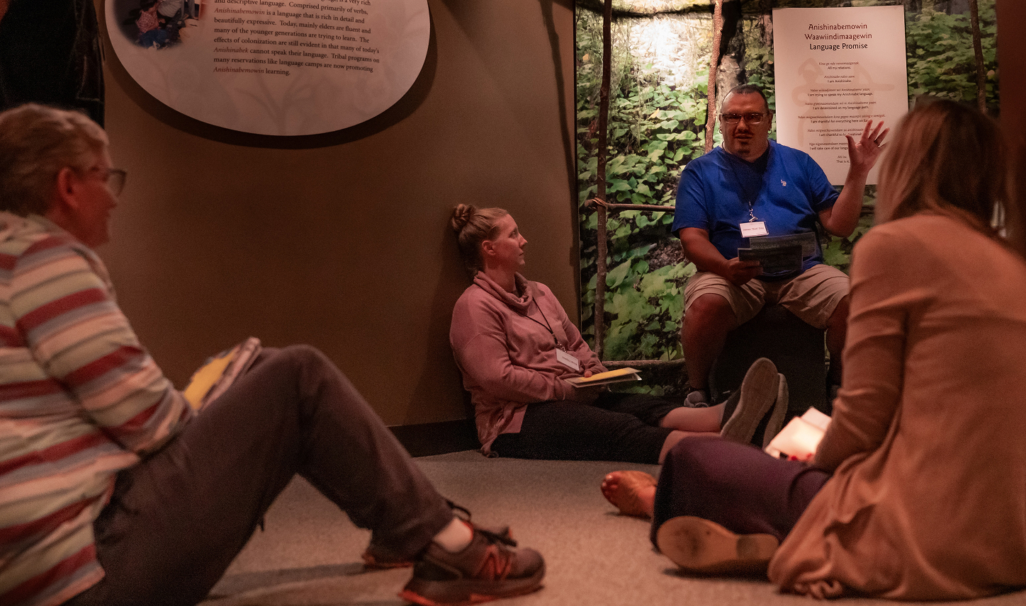 A small group of people sits in the Ziibiwing Museum listening to a facilitator talking using their hands.
