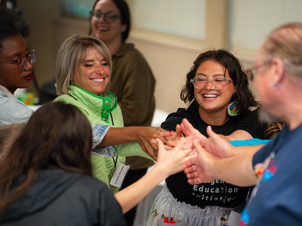 Eight people stand in a circle smiling, all reaching into the center, their hands on top of each other.